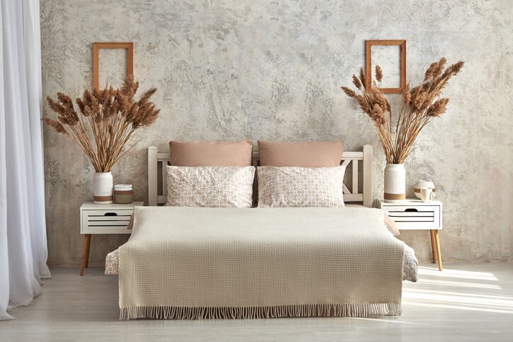 Um quarto aconchegante com uma cama bege, duas almofadas estampadas e duas mesas de cabeceira. A decoração de mesa de cabeceira apresenta vasos com capim-dos-pampas seco, enquanto um porta-retratos está pendurado na parede texturizada. A luz natural preenche o ambiente.