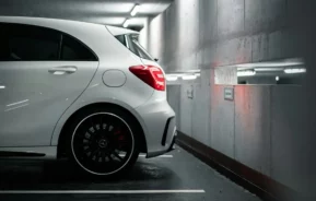Um carro branco com aros pretos está estacionado em um estacionamento coberto, ao lado de uma parede de concreto com janelas estreitas e luzes refletidas, mostrando o espaço ideal para o aluguel de garagem.