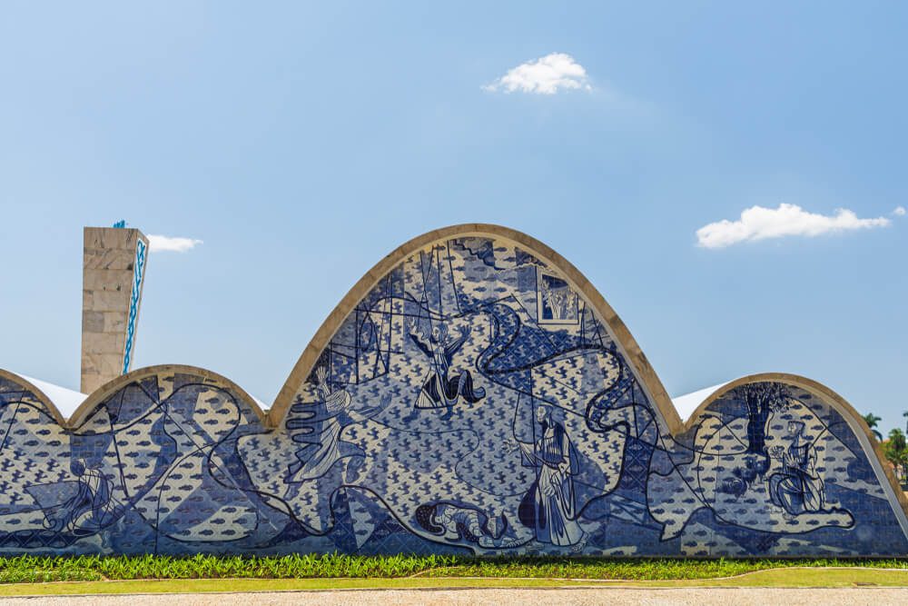 Fachada curva de edifício de concreto no Conjunto Arquitetônico da Pampulha, com um mural de azulejos azuis e brancos com pessoas, animais e padrões abstratos. Céu azul claro com nuvens dispersas ao fundo.