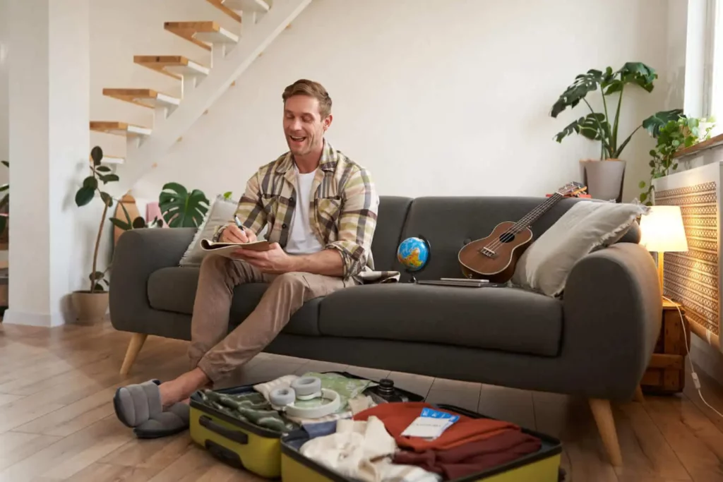 Imagem de um homem sorridente sentado em um sofá cinza escrevendo em um caderno. Uma mala, um globo, uma câmera, roupas e um ukulele estão próximos, em uma sala de estar moderna e repleta de plantas.