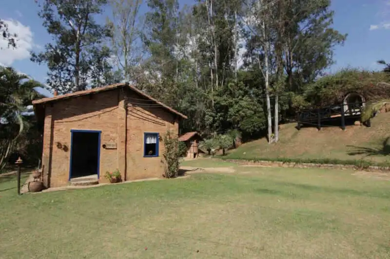 Imagem de uma pequena casa rústica de tijolos com telhado de telha no Sítio Frediani, cercada por árvores e arbustos sob um céu parcialmente nublado, para ilustrar matéria sobre o que fazer em Vinhedo em casal