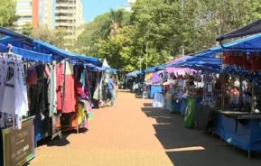 Mercado ao ar livre com marquises azuis e barracas exibindo roupas e produtos diversos em ambos os lados de uma passarela de tijolos, capturando a atmosfera vibrante da Feira Hippie de Campinas. Árvores e prédios altos se erguem ao fundo sob um céu limpo.
