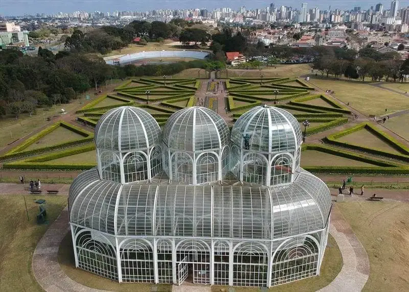 Imagem da vista aérea da famosa estufa do Jardim Botânico de Curitiba, com o jardim em estilo inglês e prédios da cidade ao fundo, para ilustrar matéria sobre o que fazer em Curitiba em 1 dia