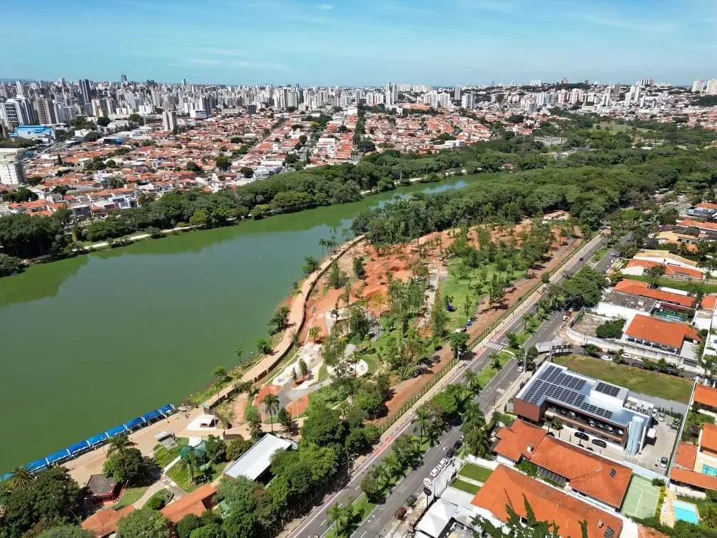 Imagem da vista aérea da Lagoa do Taquaral (Parque Portugal) em Campinas mostra um grande lago, um parque ao redor com árvores e trilhas para caminhada, residências e um denso horizonte urbano sob um céu azul e limpo.