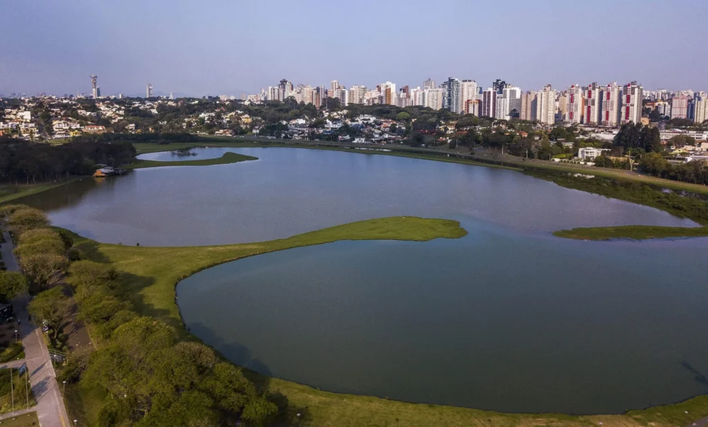 magem da vista aérea do Parque Barigui, em Curitiba, mostra lago cercado por vegetação e prédios da cidade ao fundo para ilustrar matéria sobre o que fazer em Curitiba com crianças
