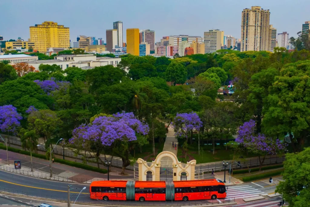 Imagem da vista aérea do Passeio Público, em Curitiba, mostra arco famoso, vegetação, ônibus vermelho e prédios ao fundo para ilustrar matéria sobre o que fazer em Curitiba com crianças