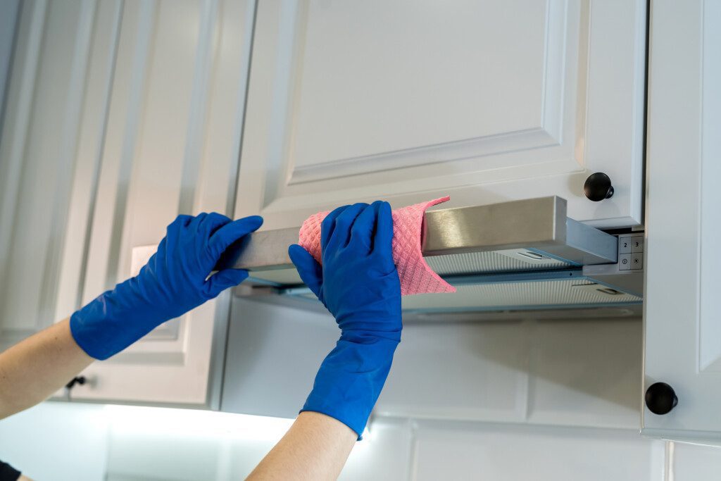 Imagem de duas mãos vestidas com luvas descartáveis azuis fazendo a limpeza de uma coifa de inox com um pano rosa