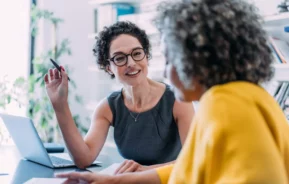 Imagem de duas mulheres sentadas ao redor de uma mesa conversando ao lado de um notebook para ilustrar matéria sobre multipropriedade