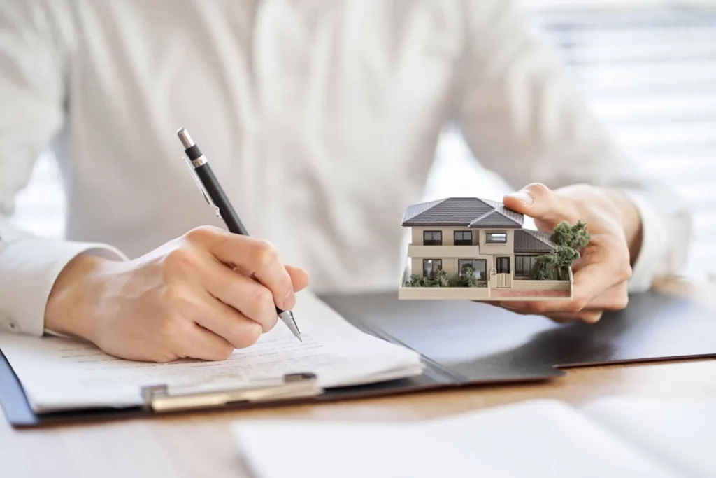 Imagem de um homem assinando um papel com uma caneta enquanto segura uma casa de madeira em tamanho miniatura com a outra mão para ilustrar matéria sobre repasse de financiamento de imóvel