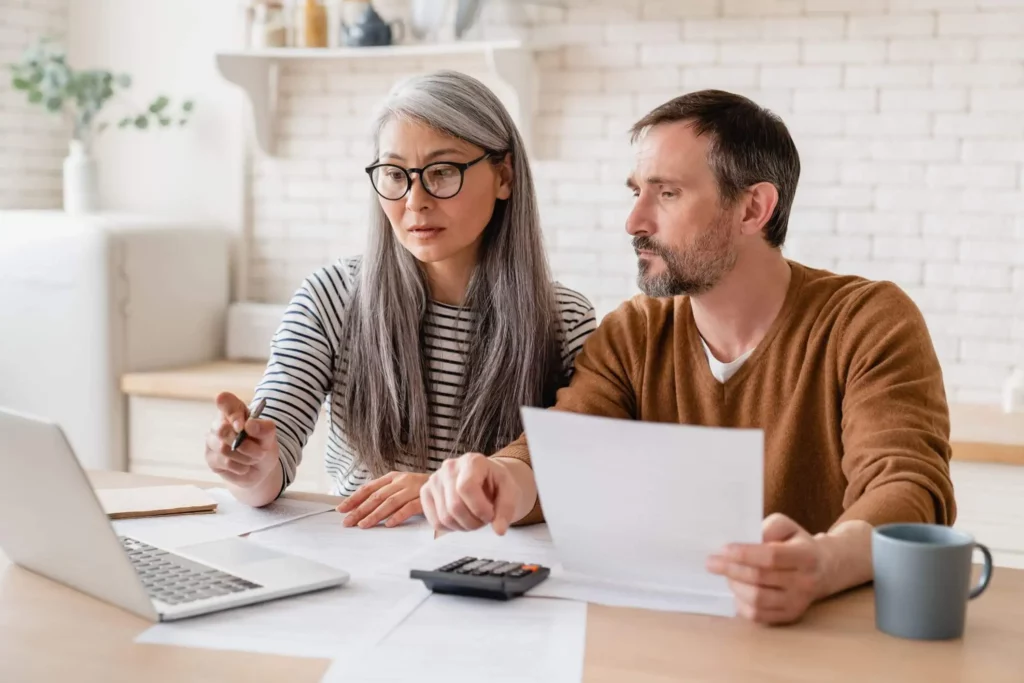 Imagem de um casal analisando alguns documentos e olhando a tela de um notebook na mesa da cozinha de casa para ilustrar matéria sobre o repasse de financiamento imobiliário