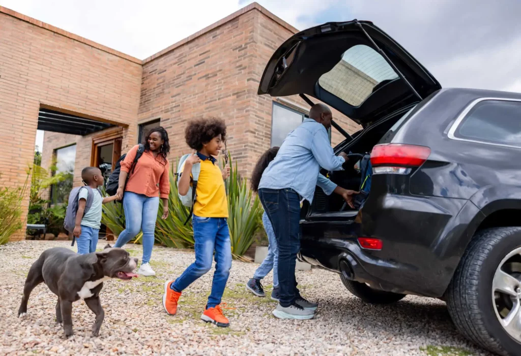 Imagem de uma família formada por um homem, uma mulher, três crianças e um cachorro tirando as malas de um carro em frente a um imóvel de alto padrão para ilustrar matéria sobre o valor de uma multipropriedade