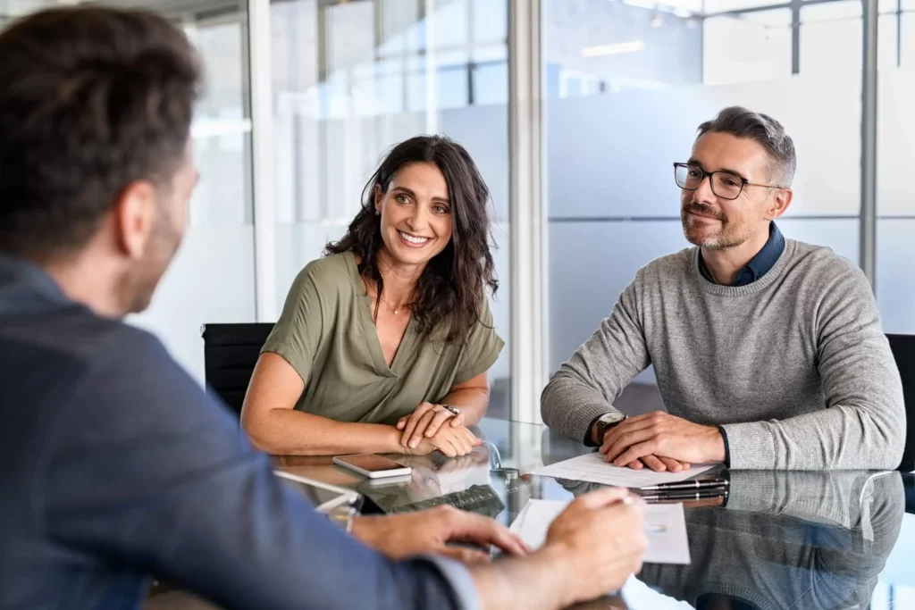 Imagem de um casal formado por um homem e uma mulher sentado em uma mesa em frente a um agente imobiliário enquanto seguram alguns papéis com as mãos para ilustrar matéria sobre: alienação fiduciária, o que significa?