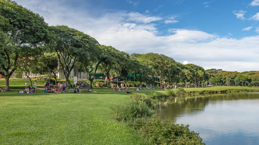 Imagem do Parque Barigui, em Curitiba, mostra lago, gramado, árvores e visitantes sentados em frente à água