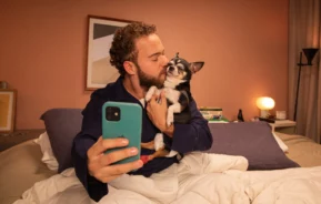 Um homem sentado na cama tira uma selfie com o celular enquanto segura e beija um pequeno cachorro preto e branco em um quarto bem iluminado — um ambiente ideal para cada caseiro, com travesseiros aconchegantes e uma luminária ao fundo.