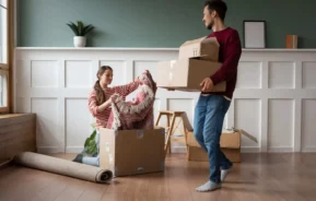 Imagem de uma mulher ajoelhada no chão enquanto desempacota um cobertor de uma caixa para ilustrar matéria sobre apartamento térreo. Do lado dela, um homem está carregando caixas de papelão em um apartamento moderno e bem iluminado, com piso de madeira e painéis brancos