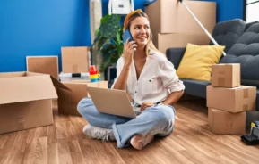 Imagem de uma mulher sorridente sentada no chão com um laptop no colo, falando ao telefone, cercada por caixas de mudança e um sofá com um travesseiro amarelo, em uma sala bem iluminada