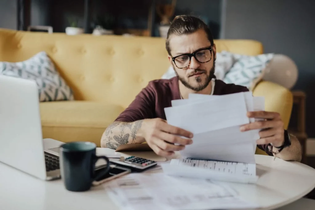 Imagem de um homem de óculos sentado em uma mesa, analisando documentos com uma expressão concentrada. Um laptop, uma calculadora, um telefone e uma caneca de café estão sobre a mesa. Um sofá amarelo e almofadas estampadas estão ao fundo