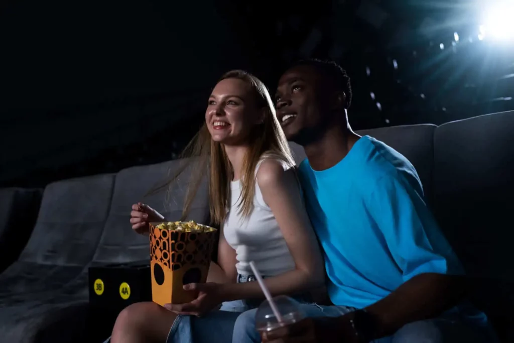 Imagem de um casal sorridente sentado junto em uma sala de cinema escura, compartilhando pipoca. A mulher veste uma blusa branca sem mangas e o homem uma camisa azul clara enquanto apreciam o filme.