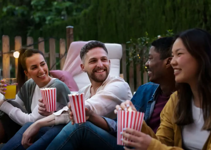 Imagem de quatro amigos sentados ao ar livre, sorrindo e conversando, cada um segurando um recipiente de pipoca listrado, para ilustrar matéria sobre os cinemas de rua em SP. Uma pessoa toma um drinque amarelo enquanto relaxam juntos, aproveitando a atmosfera casual com árvores e uma cerca de madeira atrás deles.