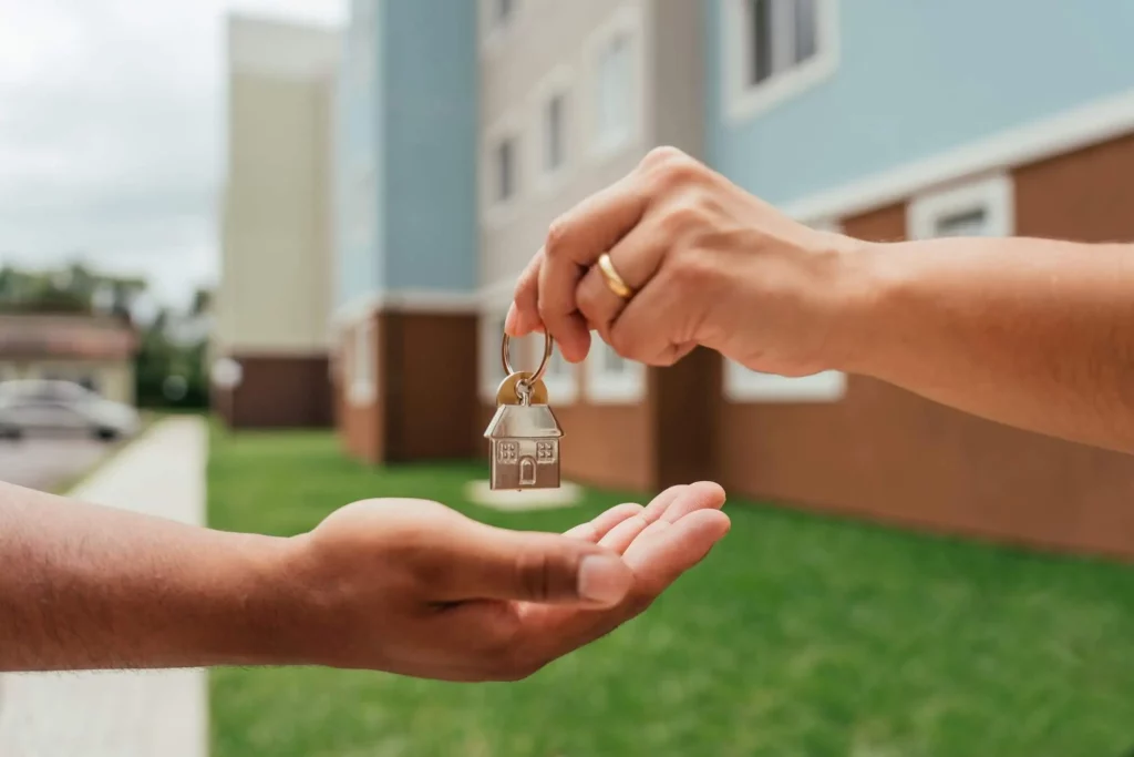 Close-up de uma pessoa entregando um chaveiro em forma de casa para outra em frente a um prédio de apartamentos