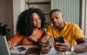 Imagem de um casal sentado em uma mesa com um caderno e um smartphone, discutindo como comprovar posse de imóvel. A mulher sorri para o homem enquanto ele escreve no caderno e verifica seu telefone. Um laptop e uma xícara de café estão sobre a mesa