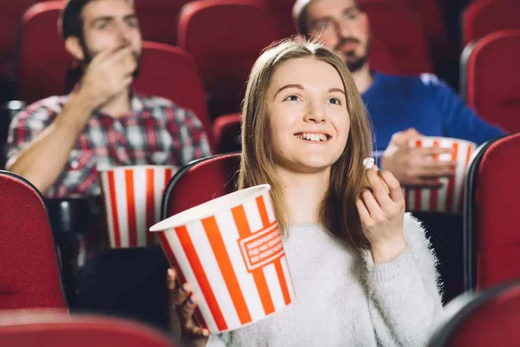 Imagem de uma jovem mulher sorrindo e comendo pipoca está sentada em um cinema com duas outras pessoas atrás dela, todas segurando potes de pipoca listrados e assistindo à tela.