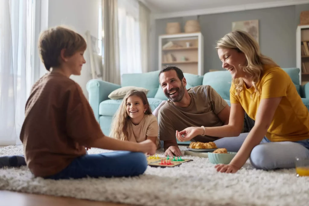 Imagem de uma família sorridente de quatro pessoas está sentada em um piso acarpetado, jogando um jogo de tabuleiro juntos em uma sala de estar aconchegante, desfrutando de um tempo de qualidade e de risadas em casa