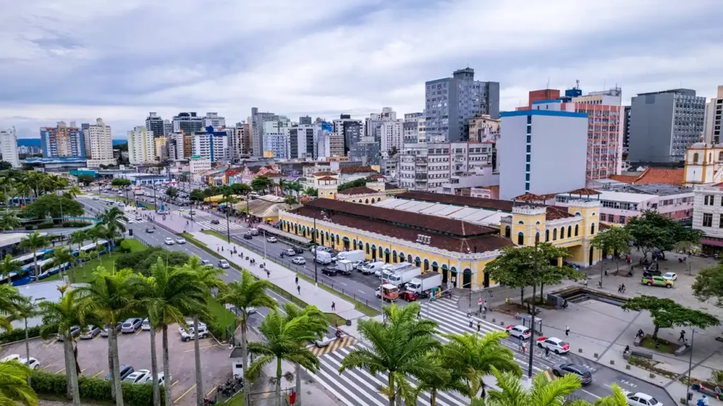 Vista aérea de uma rua movimentada da cidade, ladeada por palmeiras e tráfego, com um grande edifício histórico amarelo em frente a prédios modernos sob um céu nublado para ilustrar matéria sobre o que fazer em Florianópolis sem ser praia.