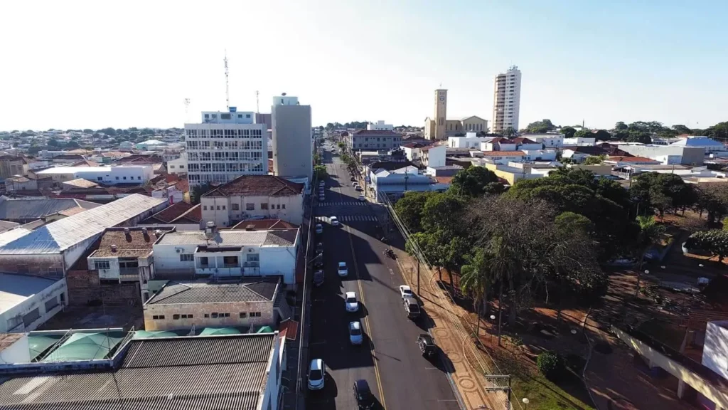 Imagem da vista aérea de uma rua de Adamantina, em SP, mostra prédios, árvores e carros estacionados sob um céu azul claro