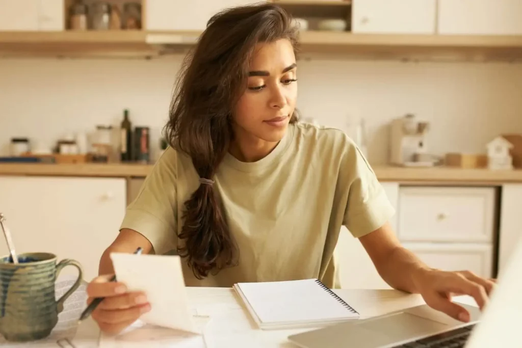 Imagem de uma mulher sentada à mesa da cozinha, olhando para um notebook enquanto segura um bloco de notas e uma caneta. Sobre a mesa há uma caneca, um caderno e alguns papéis. Ao fundo, vêem-se prateleiras e utensílios de cozinha.