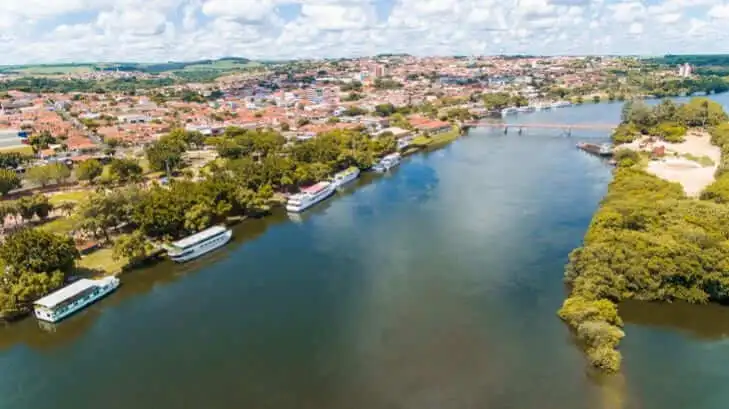 Imagem da o Rio Tietê atravessando Barra Bonita, em São Paulo, prédios de telhado vermelho, árvores verdes e vários barcos ancorados ao longo da costa sob um céu parcialmente nublado
