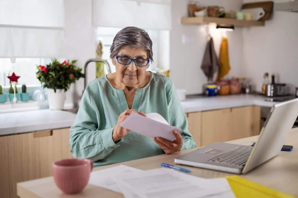 Imagem de uma senhora calculando as finanças de casa na mesa da cozinha com o auxílio de um notebook e alguns papéis para ilustrar matéria sobre como pagar IPTU atrasado