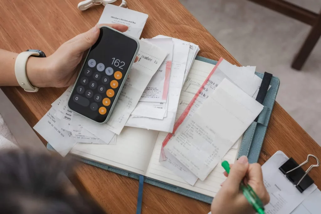 Imagem de uma pessoa gerenciando as finanças domésticas com um celular, um caderno e um lápis em cima de uma mesa de madeira para ilustrar matéria sobre como pagar o IPTU em atraso