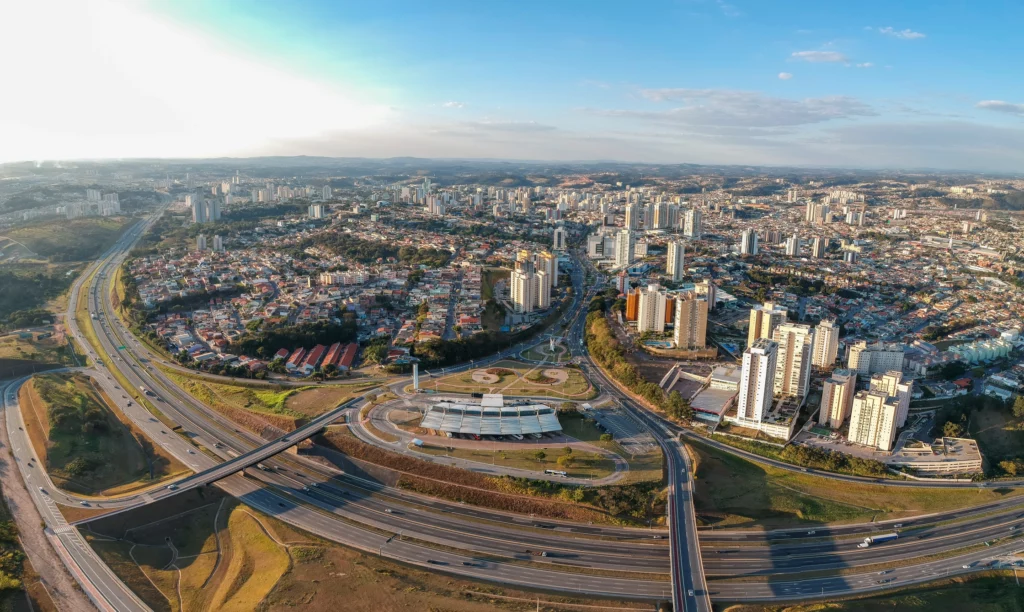 Imagem da vista aérea de Jundiaí mostra aglomerados de prédios altos, casas residenciais e rodovias que se cruzam, cercada por colinas onduladas sob um céu azul