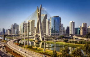 Imagem da vista aérea da ponte estaiada em SP, cercada por altos edifícios de vidro, rodovias e carros sob um céu azul claro. Árvores exuberantes margeiam a margem do rio em primeiro plano