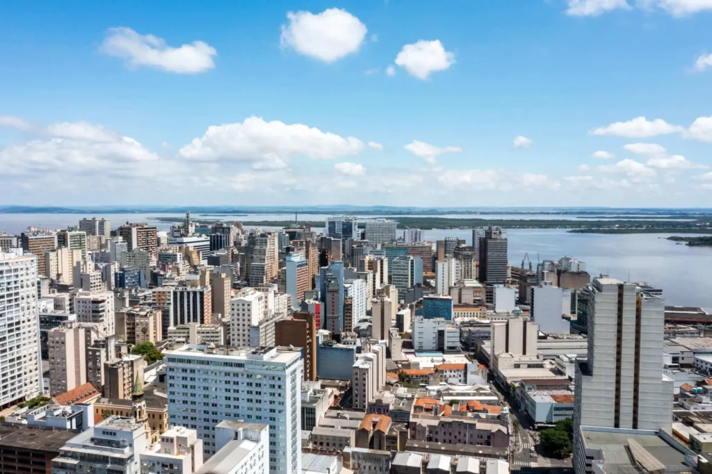 Imagem da vista aérea de Porto Alegre mostra vários prédios altos, o Rio Guaíba ao fundo e um céu azul parcialmente nublado