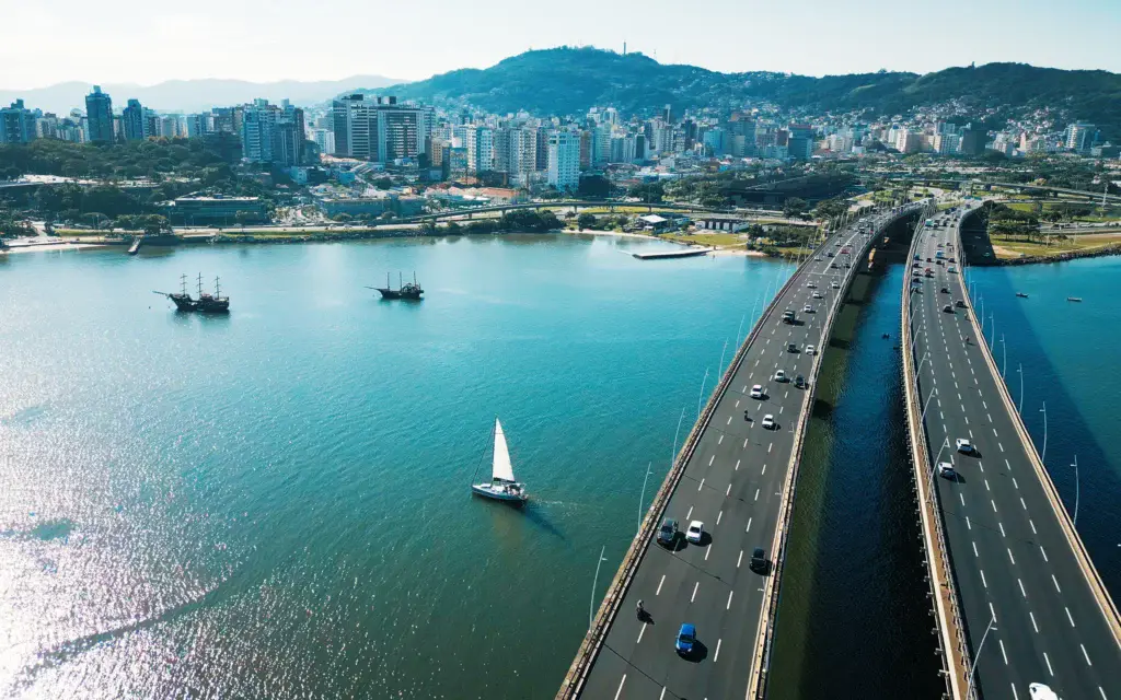 Imagem de vista aérea da cidade de Florianópolis, uma ponte com carros, veleiros na água e prédios com colinas ao fundo sob um céu claro para ilustrar matéria sobre o custo de vida em Santa Catarina, Brasil