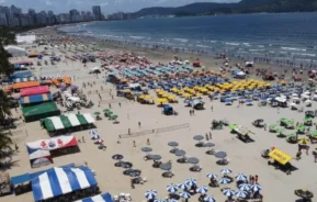 Imagem de uma praia movimentada em Santos, com muitos guarda-sóis coloridos, pessoas tomando sol, grupos jogando vôlei e barracas ao longo da areia.