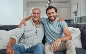 Imagem de um homem idoso com seu filho adulto, sentados um ao lado do outro em um sofá com expressão sorridente para ilustrar matéria sobre venda de imóvel para filho