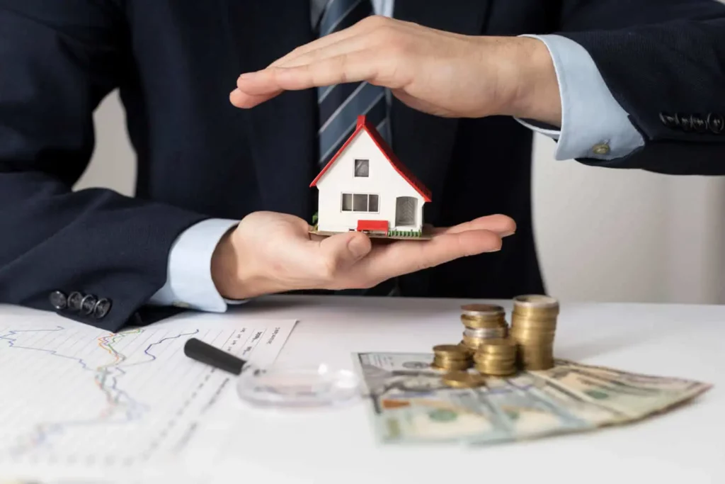 Imagem de uma pessoa de terno segurando uma pequena maquete de casa sobre uma mesa com gráficos, notas de dólar americano, moedas e uma lupa, ilustrando matéria sobre consórcio imobiliário vale a pena?