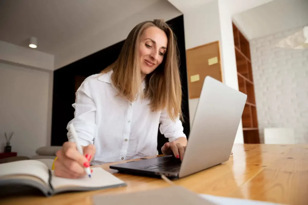 Imagem de uma mulher vestindo uma blusa branca está sentada à mesa de madeira, sorrindo enquanto pesquisa se fazer um consórcio imobiliário vale a pena, trabalhando em seu laptop e fazendo anotações em um ambiente luminoso e moderno.