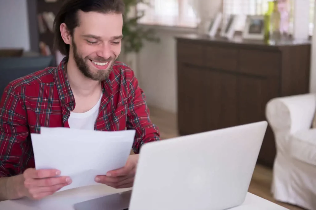 Imagem de um homem sorridente, vestindo uma camisa xadrez vermelha, está sentado à mesa, segurando alguns papéis e olhando para um laptop. A sala bem iluminada tem um armário e uma decoração elegante ao fundo.