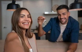 Imagem de um homem e uma mulher posando para uma foto dentro da cozinha de uma casa enquanto o homem segura as chaves de um imóvel para ilustrar matéria sobre fiador