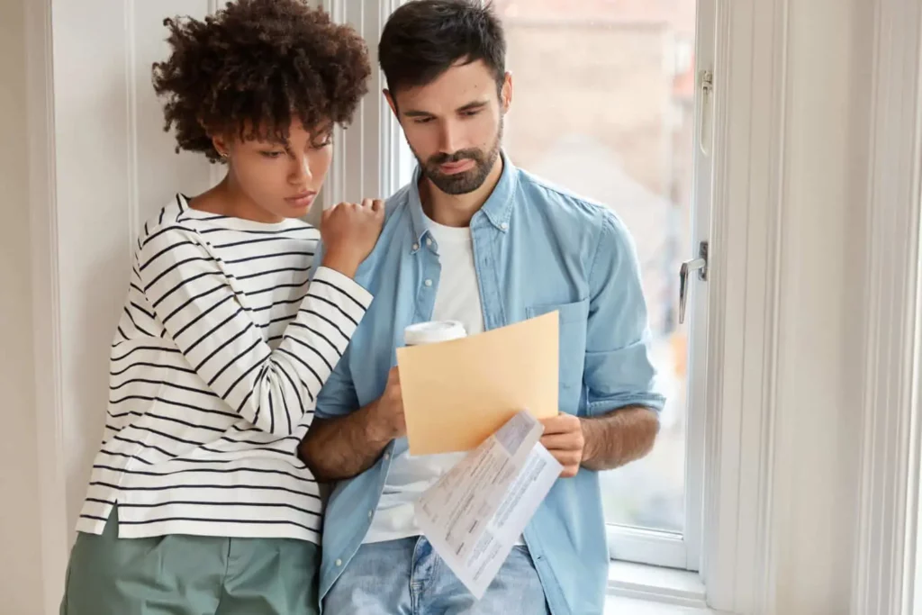Imagem de um casal formado por um homem e uma mulher encostados na janela de um imóvel avaliando as informações em um papel para ilustrar matéria sobre fiador