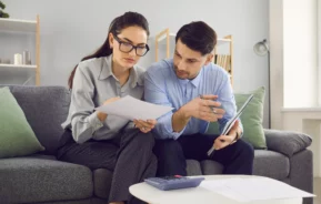 Uma mulher e um homem estão sentados em um sofá cinza, revisando documentos juntos sobre como declarar benfeitorias no imposto de renda 2025. A mulher segura papéis, enquanto o homem gesticula e segura um caderno. Uma calculadora e mais papéis estão sobre a mesa de centro branca.