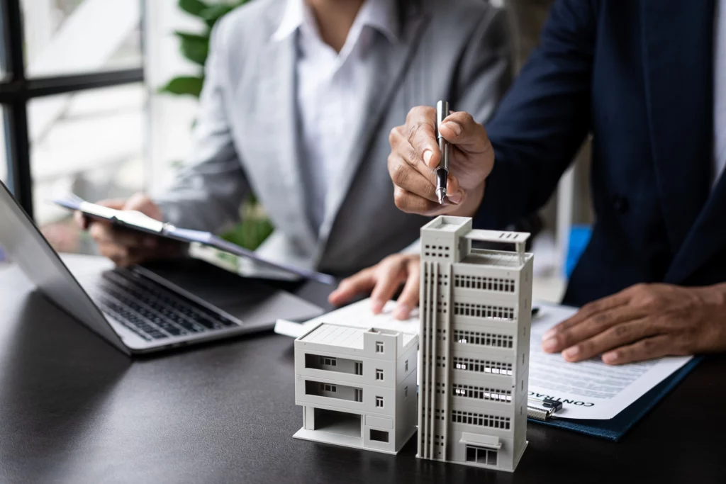 Homem de terno aponta para uma maquete de prédio enquanto um homem ao lado está com uma prancheta na mão enquanto mexe em um computador para ilustrar matéria sobre consórcio imobiliário