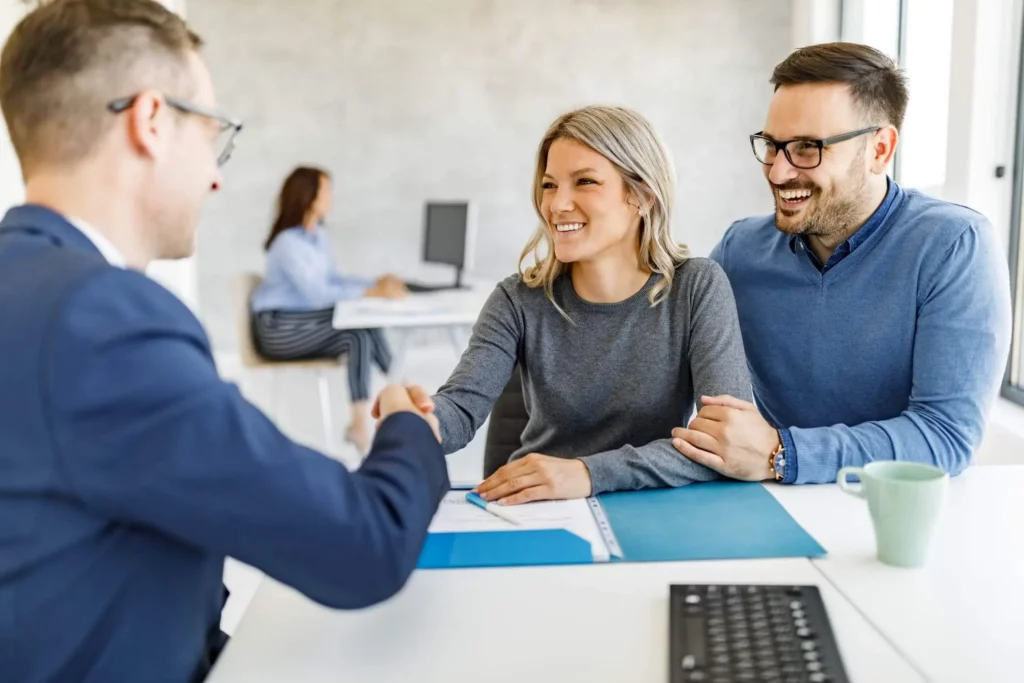 Imagem de um casal formado por um homem e uma mulher sentados em uma mesa em frente a um corretor imobiliário enquanto a mulher aperta a mão do profissional para ilustrar matéria sobre contrato de compra e venda de imóvel