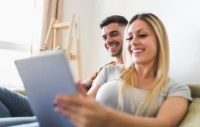 Imagem de um casal sorridente sentado junto em um sofá, olhando para a tela de um tablet para ilustrat matéria sobre o que é tabela SAC. A mulher segura o tablet enquanto o homem se senta ao lado dela. Ambos parecem relaxados e felizes em uma sala iluminada e aconchegante.