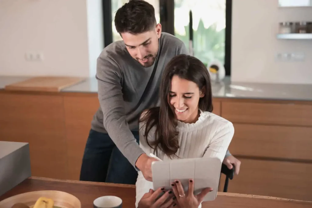 Imagem de uma mulher sorridente sentada em uma mesa olhando para um tablet, enquanto um homem está ao lado dela apontando para a tela, para ilustrar matéria sobre como funciona a Tabela SAC. Eles parecem felizes e relaxados em um ambiente de cozinha moderno.