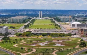 Imagem da vista aérea do Eixo Monumental de Brasília, com modernos prédios governamentais, amplos gramados verdes e a paisagem urbana ao longe sob um céu parcialmente nublado para ilustrar matéria sobre os bairros mais caros de Brasília